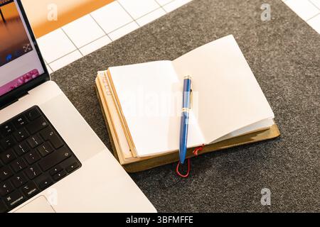 Abgewinkelte Aufnahme eines offenen leeren Notebooks und eines eleganten blauen Stiftes mit goldenem Besatz, bereit für den Einsatz neben einem modernen Laptop auf einem gut organisierten Schreibtisch. Diese KOM Stockfoto