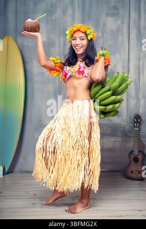 Ethnische fröhliche Frau in traditioneller hawaiianischer Tracht mit Bananen und einem Kokosdrink in einem älteren Studio mit Requisiten Stockfoto