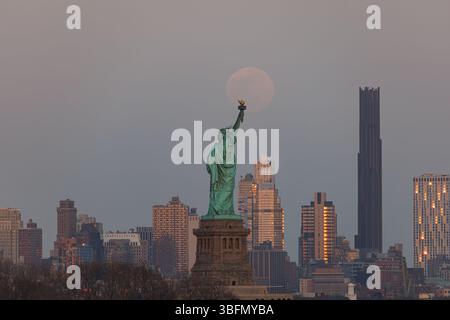 Die Freiheitsstatue steht in der Abenddämmerung hoch, während der Vollmond dahinter aufsteigt und ein magisches Leuchten über die Skyline von New York City wirft. Stockfoto