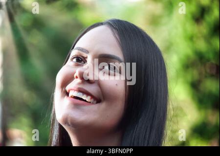 Porträt einer fröhlichen jungen Frau mit langen, dunklen Haaren, die lächelnd und aufblickend sind. Der Hintergrund ist mit sanftem Grün gefüllt, was dem Bild ein gutes Bild verleiht Stockfoto