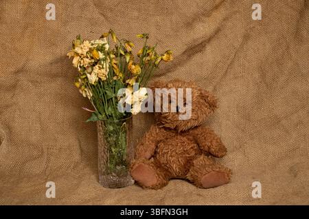 Teddybär neben einer Vase getrockneter Blumen auf einem Sackleinen Hintergrund, ein Symbol für Traurigkeit, Verlust und Abschied Stockfoto