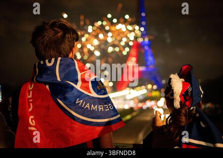 Ein Paris Saint-Germain Fan, der in eine PSG-Flagge gehüllt ist, sieht ein Feuerwerk am Eiffelturm. Stockfoto