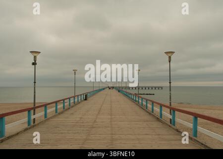 Symmetrische Perspektive einer langen Holzpier, die sich unter bewölktem Himmel in die Ostsee erstreckt, mit modernen Straßenlaternen an beiden Seiten. Stockfoto