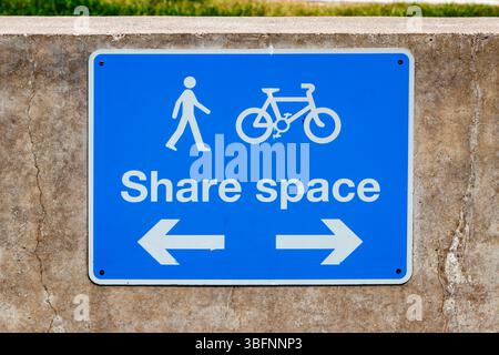Blaues Schild, das einen gemeinsamen Raum für Fußgänger und Radfahrer auf einer Betonwand mit Richtpfeilen darunter anzeigt Stockfoto