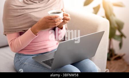 Online-Arbeit, Bildung, Kommunikation. Unkenntlich schwarze Frau in Hijab mit Laptop und trinken Kaffee zu Hause Stockfoto
