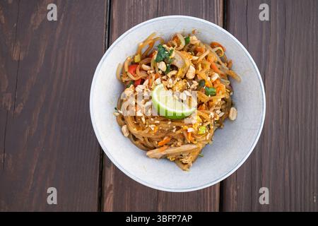 Eine verführerische Schüssel Pad See Ew mit breiten Reisnudeln, Hühnchen, Gemüse und einem frischen Limettenkeil auf Holz. Stockfoto