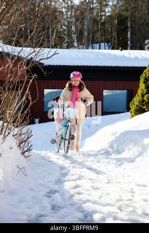 Tween geht mit ihrem Fahrrad nach Hause an einem sonnigen Wintertag in einem finnischen Vorort. Schnee auf dem Boden, ein echter Moment des nordischen Alltags. Stockfoto