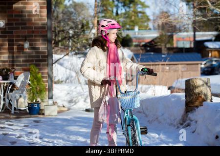 Tween geht mit ihrem Fahrrad nach Hause an einem sonnigen Wintertag in einem finnischen Vorort. Schnee auf dem Boden, ein echter Moment des nordischen Alltags. Stockfoto