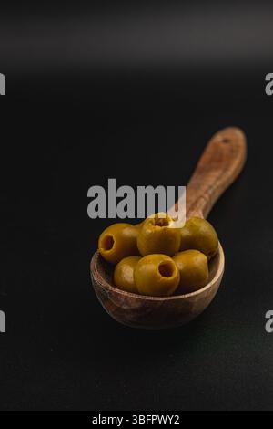Ein Holzlöffel fasst mehrere grüne Oliven und zeigt ihre glatte Textur und satte Farbe vor einem kontrastierenden dunklen Hintergrund. Die Einfachheit des Stockfoto