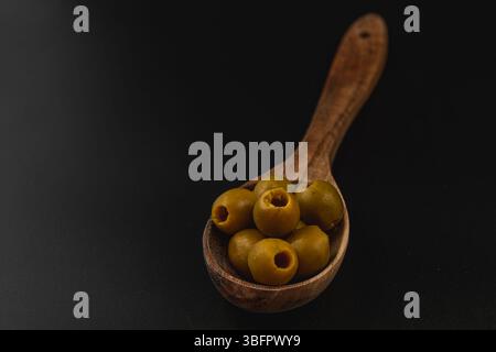 Ein Holzlöffel fasst mehrere frische grüne Oliven und zeigt ihre glatte Textur und einzigartige Form. Der kontrastfarbene dunkle Hintergrund verstärkt die Appeti Stockfoto