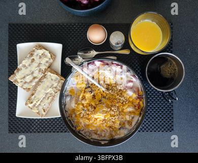 Berlin, Deutschland. Juni 2025. Eine Schüssel Joghurt mit Erdbeeren, einer gehackten Banane, ungesüßten Cornflakes und Haferflocken steht vor dem Frühstück neben einem Teller mit zwei Scheiben Crispbrot mit Butter, einem gekochten Ei, einem Glas Orangensaft und einer Tasse schwarzen Kaffee. Quelle: Soeren Stache/dpa/Alamy Live News Stockfoto