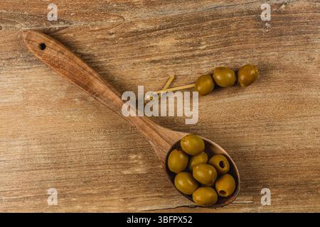 Ein Holzlöffel hält mehrere grüne Oliven, während ein Spieß mit Oliven auf dem Holztisch liegt. Die natürliche Textur des Holzes verleiht dem Set Wärme Stockfoto