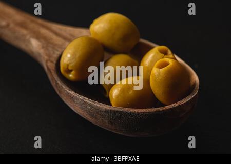 Ein rustikaler Holzlöffel fasst mehrere grüne Oliven und zeigt ihre glänzende Textur und leuchtende Farbe vor dunklem Hintergrund. Die Komposition ist hochgradig Stockfoto