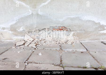 Beschädigte Betonwand mit abblätterndem Putz und sichtbaren roten Ziegeln, umgeben von umgestürzten Trümmern und verwitterten Pflasterziegeln. Stockfoto