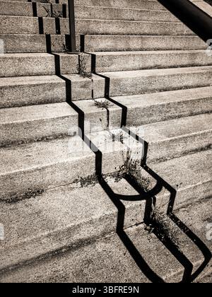 Unebene Betonstufen in einem städtischen Gebiet weisen Risse auf, die mit Unkraut gefüllt sind, das in den Spalten wächst. Schatten eines Treppengeländes Stockfoto