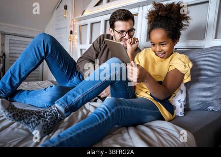 Alleinerziehender Vater spielt mit seinem multiethnischen, vielfältigen Kind auf dem Bett mit einem digitalen Tablet zu Hause Stockfoto
