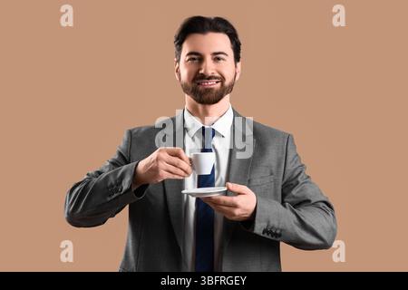 Lächelnder, gutaussehender junger Mann im Anzug mit einer Tasse Kaffee auf beigefarbenem Hintergrund Stockfoto