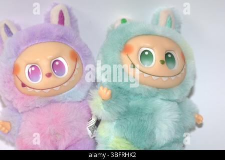 Zwei verspielte Labubu-Spielzeugfiguren in flauschigen grünen und rosa Monsterkostümen und einem schelmischen Lächeln, die vor weißem Hintergrund aufgestellt werden. Stockfoto