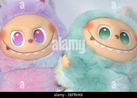 Zwei verspielte Labubu-Spielzeugfiguren in flauschigen grünen und rosa Monsterkostümen und einem schelmischen Lächeln, die vor weißem Hintergrund aufgestellt werden. Stockfoto