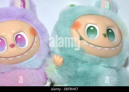 Zwei verspielte Labubu-Spielzeugfiguren in flauschigen grünen und rosa Monsterkostümen und einem schelmischen Lächeln, die vor weißem Hintergrund aufgestellt werden. Stockfoto