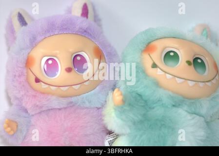 Zwei verspielte Labubu-Spielzeugfiguren in flauschigen grünen und rosa Monsterkostümen und einem schelmischen Lächeln, die vor weißem Hintergrund aufgestellt werden. Stockfoto