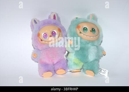 Zwei verspielte Labubu-Spielzeugfiguren in flauschigen grünen und rosa Monsterkostümen und einem schelmischen Lächeln, die vor weißem Hintergrund aufgestellt werden. Stockfoto