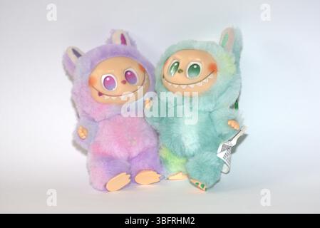 Zwei verspielte Labubu-Spielzeugfiguren in flauschigen grünen und rosa Monsterkostümen und einem schelmischen Lächeln, die vor weißem Hintergrund aufgestellt werden. Stockfoto
