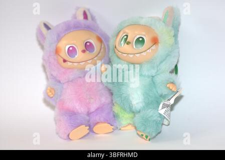 Zwei verspielte Labubu-Spielzeugfiguren in flauschigen grünen und rosa Monsterkostümen und einem schelmischen Lächeln, die vor weißem Hintergrund aufgestellt werden. Stockfoto