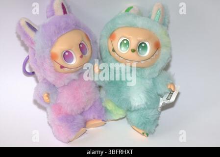 Zwei verspielte Labubu-Spielzeugfiguren in flauschigen grünen und rosa Monsterkostümen und einem schelmischen Lächeln, die vor weißem Hintergrund aufgestellt werden. Stockfoto