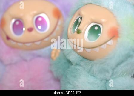 Zwei verspielte Labubu-Spielzeugfiguren in flauschigen grünen und rosa Monsterkostümen und einem schelmischen Lächeln, die vor weißem Hintergrund aufgestellt werden. Stockfoto