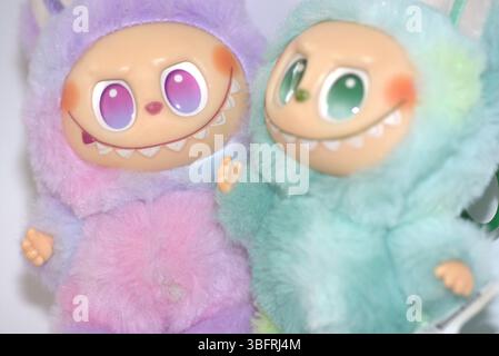 Zwei verspielte Labubu-Spielzeugfiguren in flauschigen grünen und rosa Monsterkostümen und einem schelmischen Lächeln, die vor weißem Hintergrund aufgestellt werden. Stockfoto