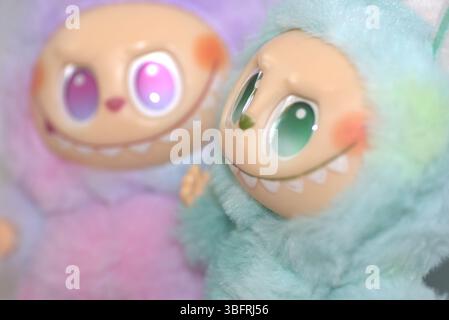 Zwei verspielte Labubu-Spielzeugfiguren in flauschigen grünen und rosa Monsterkostümen und einem schelmischen Lächeln, die vor weißem Hintergrund aufgestellt werden. Stockfoto