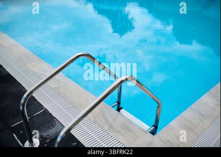 Nahaufnahme einer Edelstahlleiter, die in einen sauberen blauen Swimmingpool führt. Reflexionen von Wolken auf der Wasseroberfläche. Ruhiges Resort Stockfoto
