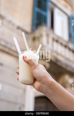 Nahaufnahme einer Hand, die Zitronengranita mit Strohhalmen gegen verschwommenes italienisches Gebäude hält Stockfoto