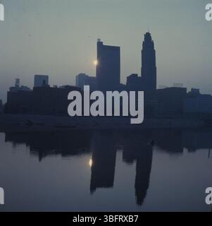Ein ruhiger Morgenblick auf die Skyline von Columbus, mit einem Kanu, das bei Sonnenaufgang auf dem Scioto River driftet und ein goldenes Licht auf die Gebäude der Stadt wirft. Stockfoto