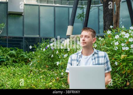 Junger Mann mit Laptop, der im Stadtpark mit grünen Pflanzen und Blumen sitzt, eine nachdenkliche Pause von der Arbeit macht, die Natur und die frische Luft in moderner Atmosphäre genießt Stockfoto