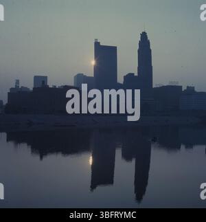 Ein Kanu gleitet entlang des Scioto River, während die Sonne über der Skyline von Columbus aufgeht und den Himmel in sanften Orange- und Rosa-Tönen malt, während warme Reflexe auf dem Wasser reflektiert werden. Stockfoto
