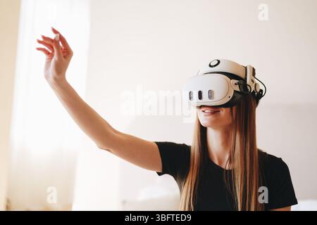 Die junge Frau erlebt virtuelle Realität in einem gemütlichen Innenbereich, während sie lächelt und mit digitalen Elementen interagiert Stockfoto