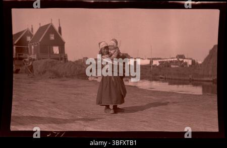 Dieses Foto aus dem Jahr 1913 zeigt ein niederländisches Mädchen in traditioneller Kleidung, das während der Heuernte in Marken, Niederlande, ein Baby hält. Die Arrases nahmen das alltägliche Leben während ihrer Europafahrten auf und dokumentierten ländliche Szenen. Stockfoto