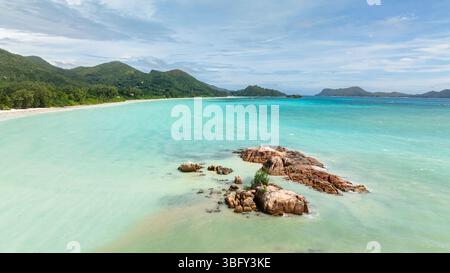 Sanfte Wellen umgeben felsige Ausläufer im ruhigen türkisfarbenen Wasser, mit üppigen Hügeln und einer Sandküste in der Ferne. Anse Volbert Beach. Praslin, Seychellen. Stockfoto