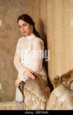 Eine junge Frau in einem Vintage-Kleid lehnt sich anmutig an einen kunstvollen Stuhl und strahlt Eleganz aus. Stockfoto