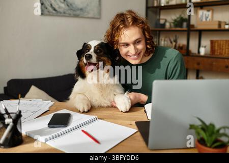 Ein junger Mann sitzt an einem Holztisch und lächelt, während er online mit seinem Hund interagiert. Stockfoto