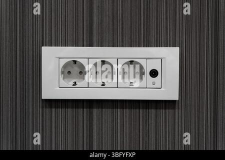 Nahaufnahme einer dreifachen Steckdose im europäischen Stil, die an einer dunklen, vertikal gestreiften, strukturierten Wand installiert ist. Stockfoto
