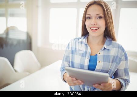 Büroangestellter, der sich neben Tisch und Stühlen durch eine Glaswand beugt und ein Tablet unter großen Fenstern hält Stockfoto
