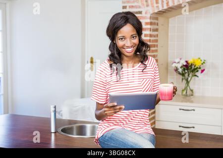 Afroamerikanische Frau, die auf hölzerner Arbeitsplatte in der Küche sitzt und Tablette und rosa Kaffeetasse hält Stockfoto
