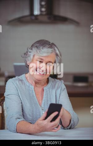 Seniorin hält Smartphone und lächelt zu Hause Küchentisch mit Laptop, Motorhaubenbelüftung, Gegenständen Stockfoto