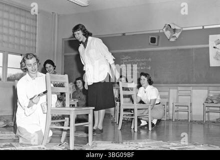 Foto von A.V. Shirk aus dem Jahr 1955, das einen Lehrer zeigt, der mit Schülern Stühle bemalt, möglicherweise als Teil einer Hauswirtschaftsklasse oder Kunstklasse. Dieses Bild spiegelt Shirks frühe fotografische Arbeit wider und erfasst Momente des Alltags in Ohio. Stockfoto