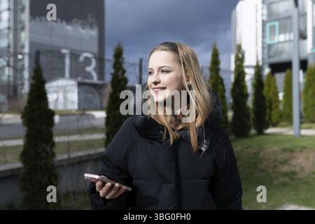 Eine junge Frau steht draußen und lächelt, während sie ihr Smartphone benutzt. Sie ist umgeben von modernen architektonischen Gebäuden und Grün unter einem bewölkten Himmel Stockfoto