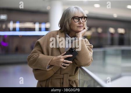 Eine Frau mit stilvoller Brille lehnt sich an ein Geländer und hält nachdenklich ihr Smartphone. Sie scheint sich in einer zeitgenössischen Umgebung verloren zu haben Stockfoto