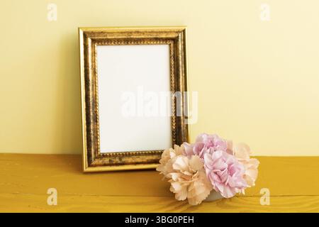 Leerer Bilderrahmen mit Nelkenblumen auf Holztisch. Gelber Hintergrund Stockfoto